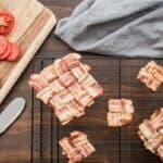 Bacon weaves on a cooling rack with sliced tomatoes, lettuce, bread, and a butter knife on a cutting board. Nearby is a gray cloth.