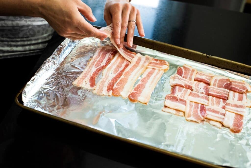 Hands arranging bacon strips into a weave pattern on a foil-lined baking sheet.