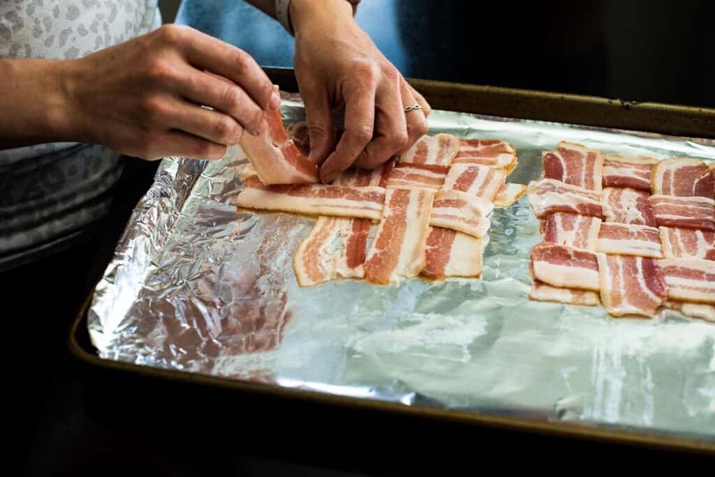 Hands weave bacon strips into a lattice pattern on a foil-lined baking sheet.