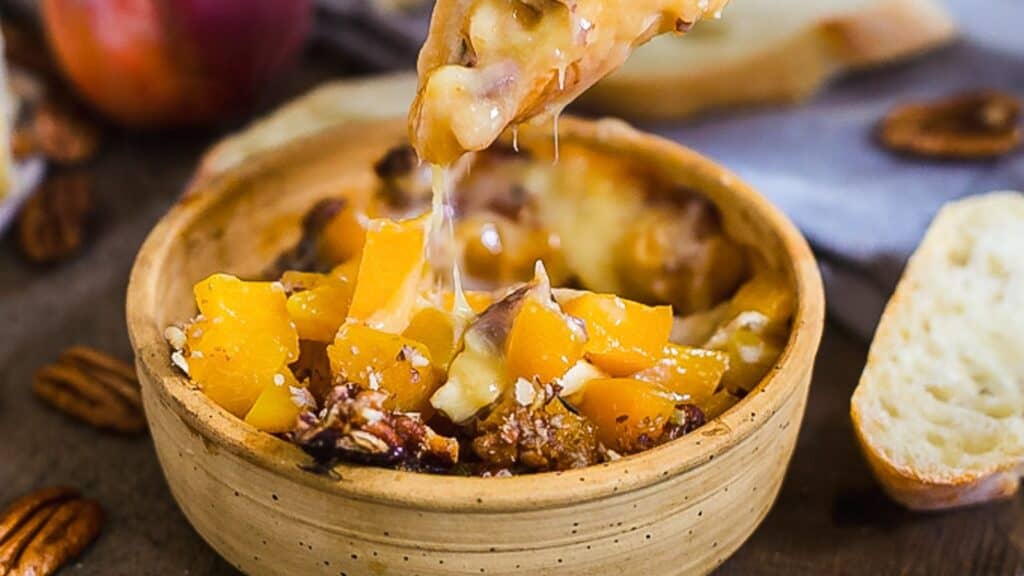 A wooden bowl of melted cheese and chopped peaches with scattered nuts. Bread slices are visible in the background.