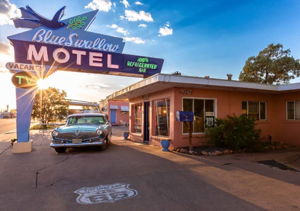 Vintage car parked at Blue Swallow Motel with retro neon sign. Sun setting, casting warm light. Route 66 emblem painted on the ground.