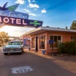 Vintage car parked at Blue Swallow Motel with retro neon sign. Sun setting, casting warm light. Route 66 emblem painted on the ground.