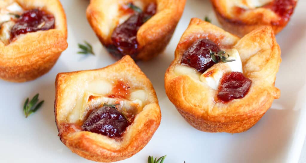 Bite-sized pastry cups filled with melted cheese, topped with cranberry sauce and garnished with fresh thyme on a white surface.