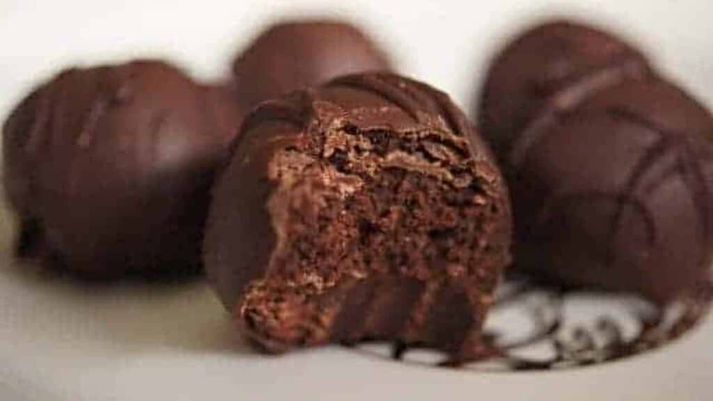 Close-up of chocolate truffles, with one having a bite taken out, perfect for New Year's Eve desserts. They sit elegantly on a white plate with a decorative drizzle.