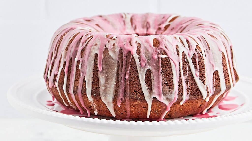 A bundt cake with pink and white icing drizzles sits on a white pedestal cake stand.