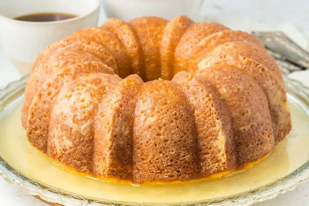 A bundt cake sits on a glass plate, covered in a light glaze. A cup of coffee is blurred in the background.