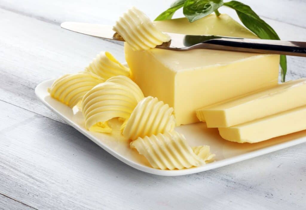 A plate of fancy butter features several elegant curls and slices. A knife rests gracefully on the butter block, accompanied by a green leaf garnish on top.