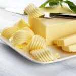 A plate of fancy butter features several elegant curls and slices. A knife rests gracefully on the butter block, accompanied by a green leaf garnish on top.