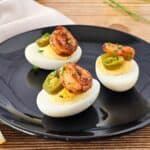 Three deviled eggs topped with shrimp and jalapeño slices on a black plate, placed on a wooden table with a lemon wedge nearby.