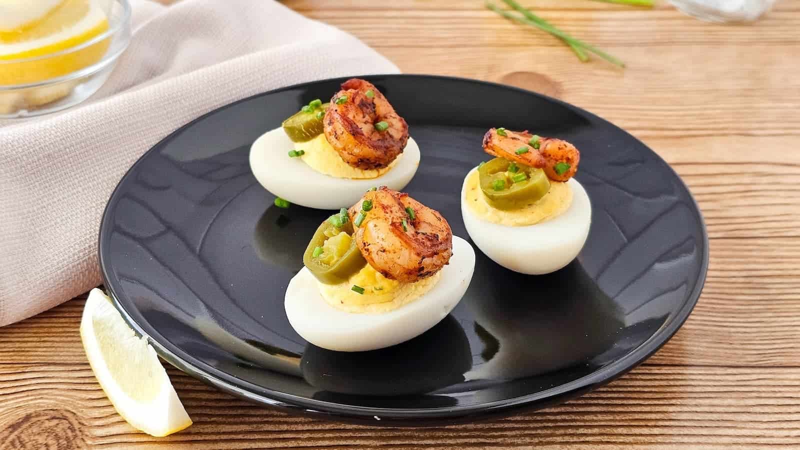 Three deviled eggs topped with shrimp and jalapeño slices on a black plate, placed on a wooden table with a lemon wedge nearby.