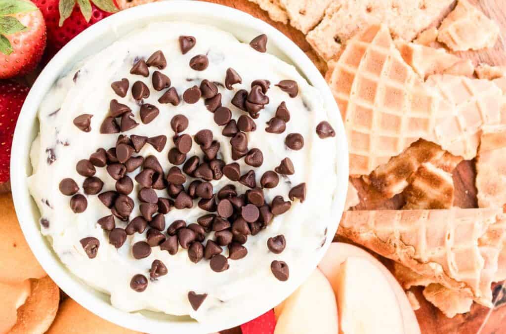 A bowl of cream cheese dip with chocolate chips, surrounded by waffle pieces, cookies, strawberries, and apple slices.