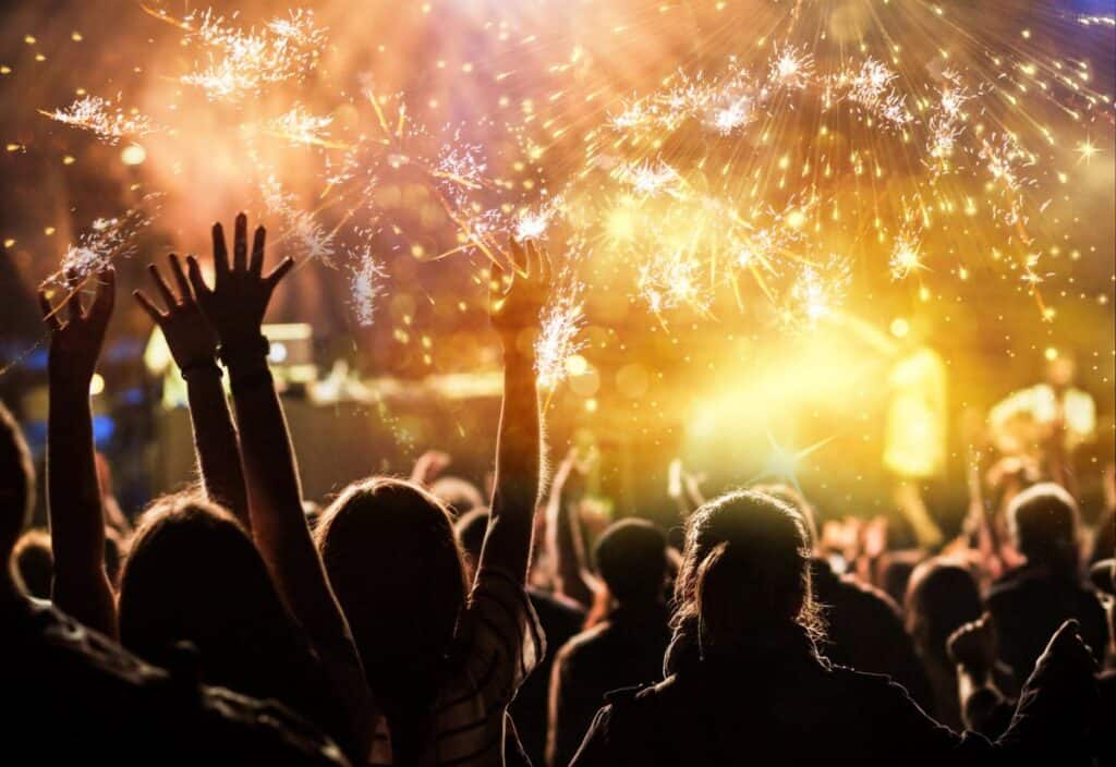 A crowd of people with raised arms enjoys a vibrant fireworks display at night.