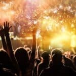 A crowd of people with raised arms enjoys a vibrant fireworks display at night.