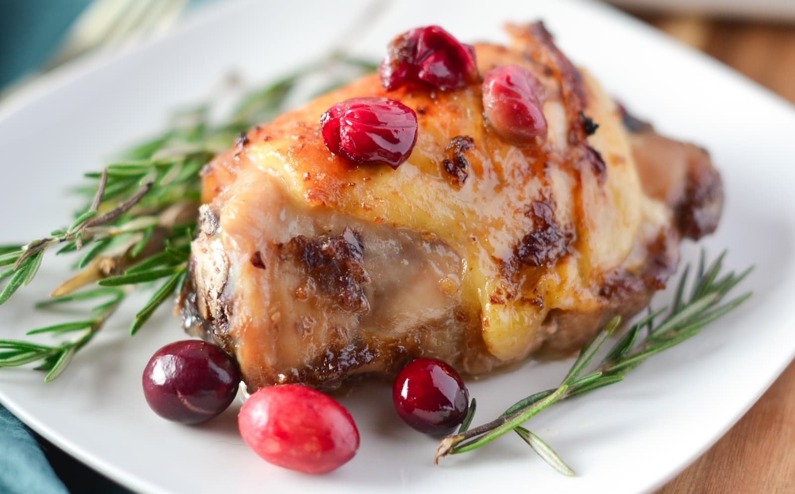 A piece of roasted chicken thighs with cranberry sauce, garnished with rosemary, rests elegantly on a white plate.