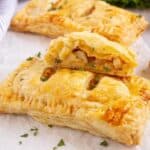 Rectangular puff pastries filled with diced chicken and herbs, displayed on parchment paper, with one pastry cut open to show the filling. A striped cloth is visible on the side.