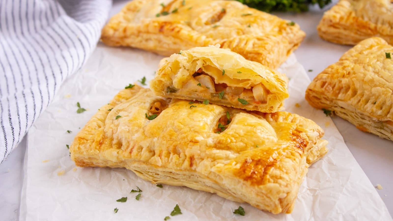 Golden-brown puff pastry pockets filled with a savory mixture of chicken pot pie, placed on white parchment paper, with a striped cloth and green herbs in the background.