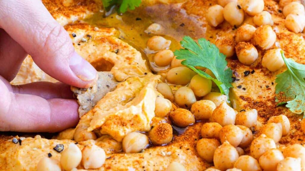 Close-up of a hand dipping a piece of bread into a dish of hummus, topped with whole chickpeas, olive oil, paprika, and garnished with fresh parsley.