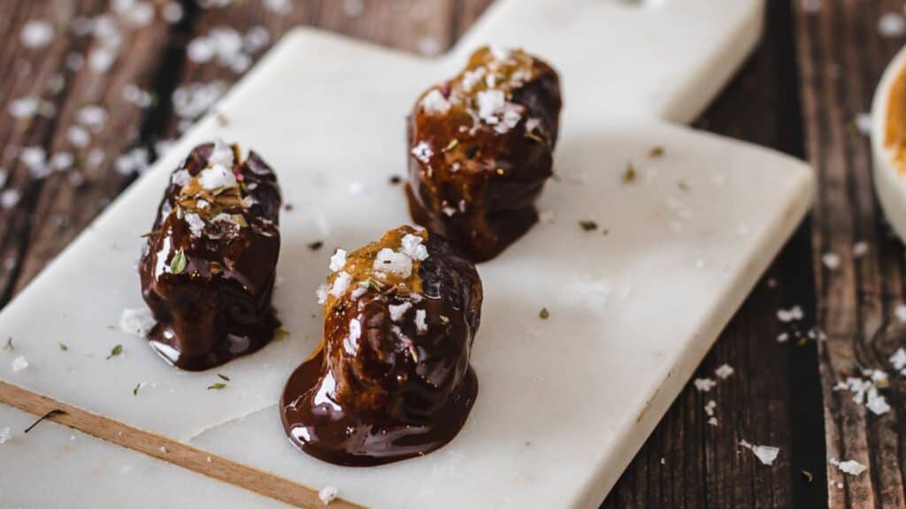 Three chocolate-covered dates topped with salt and herbs on a marble cutting board.