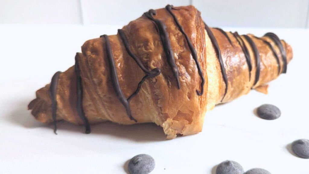 A croissant drizzled with chocolate sits on a white surface, surrounded by a few chocolate chips.