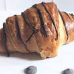 A croissant drizzled with chocolate sits on a white surface, surrounded by a few chocolate chips.