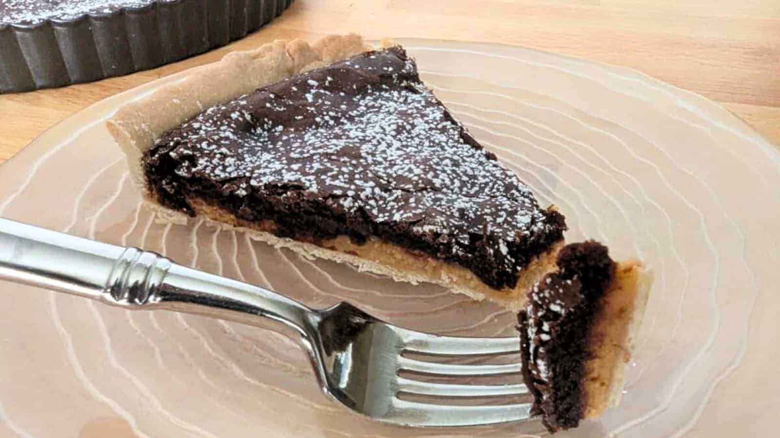 A slice of chocolate tart on a clear plate with a fork. The tart is topped with powdered sugar.