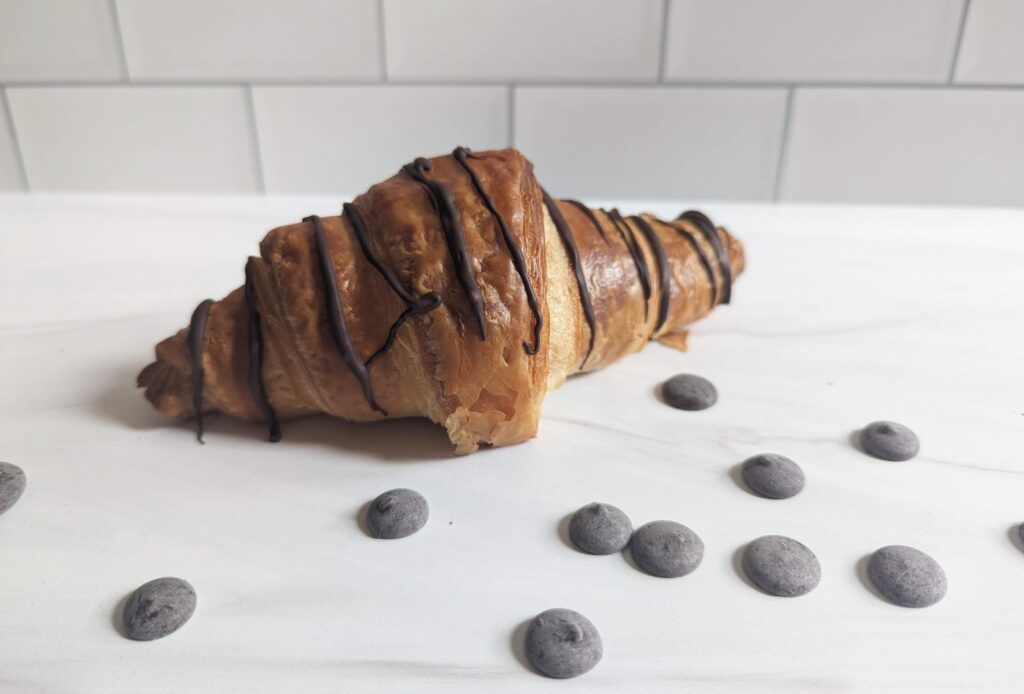 Chocolate-drizzled croissant on a white surface, surrounded by small chocolate pieces.