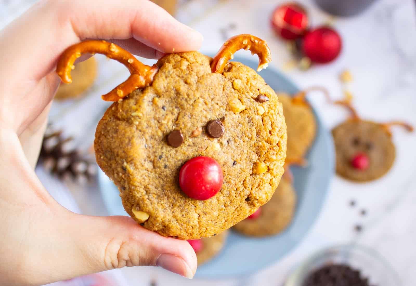 A plate of Christmas reindeer cookies with pretzel antlers, chocolate chip eyes, and red M&M noses, surrounded by holiday decorations and baking ingredients.