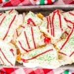 Festive holiday bark with white frosting, red drizzle, and green sprinkles on a tray, set on a red and white checkered cloth.