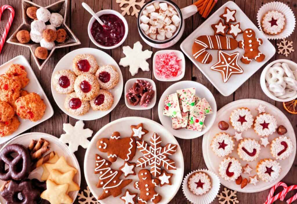 Assorted holiday cookies and sweets on a wooden table, featuring allergy-friendly baking delights like gingerbread, snowflake cookies, chocolate treats, and a mug of hot chocolate topped with marshmallows.