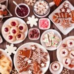 Assorted holiday cookies and sweets on a wooden table, featuring allergy-friendly baking delights like gingerbread, snowflake cookies, chocolate treats, and a mug of hot chocolate topped with marshmallows.