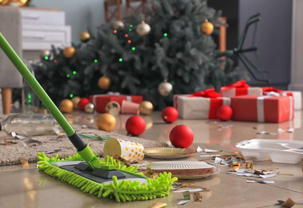 A green mop cleans a floor littered with confetti, paper plates, cups, and Christmas decorations. A decorated Christmas tree and wrapped gifts are in the background.