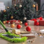 A green mop cleans a floor littered with confetti, paper plates, cups, and Christmas decorations. A decorated Christmas tree and wrapped gifts are in the background.