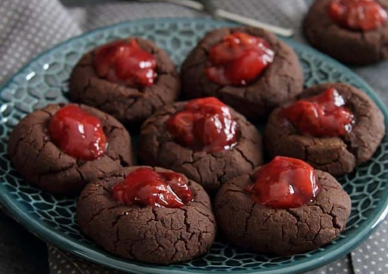 A blue plate holds eight chocolate thumbprint cookies filled with red cherry topping.