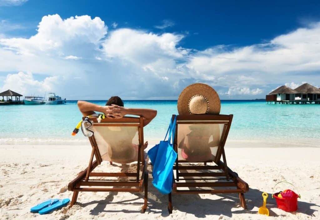 Two people relax on beach chairs facing a clear turquoise sea, under a blue sky. A blue bag and beach toys are on the sand beside them. Nearby are overwater bungalows.