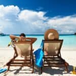 Two people relax on beach chairs facing a clear turquoise sea, under a blue sky. A blue bag and beach toys are on the sand beside them. Nearby are overwater bungalows.