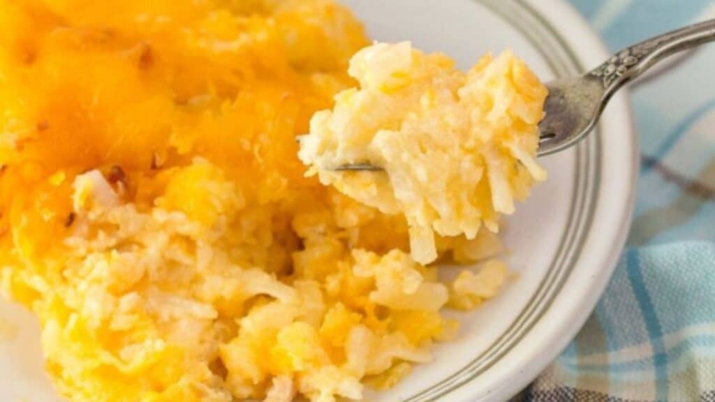 A plate of cheesy hash brown casserole with a forkful shown, featuring a golden, crispy top on a white plate—an easy crockpot creation that delights.