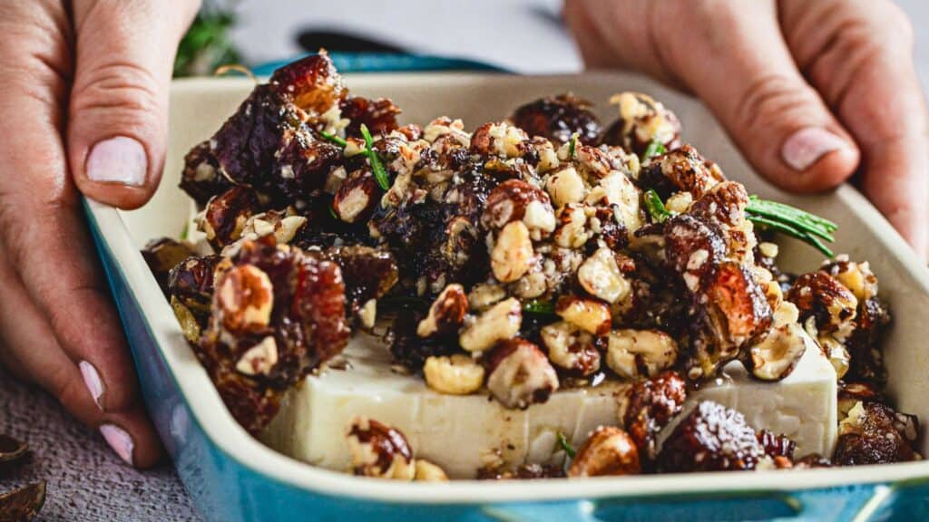 A dish with a block of cheese topped with nuts, herbs, and dried fruits in a ceramic dish being held by a person.