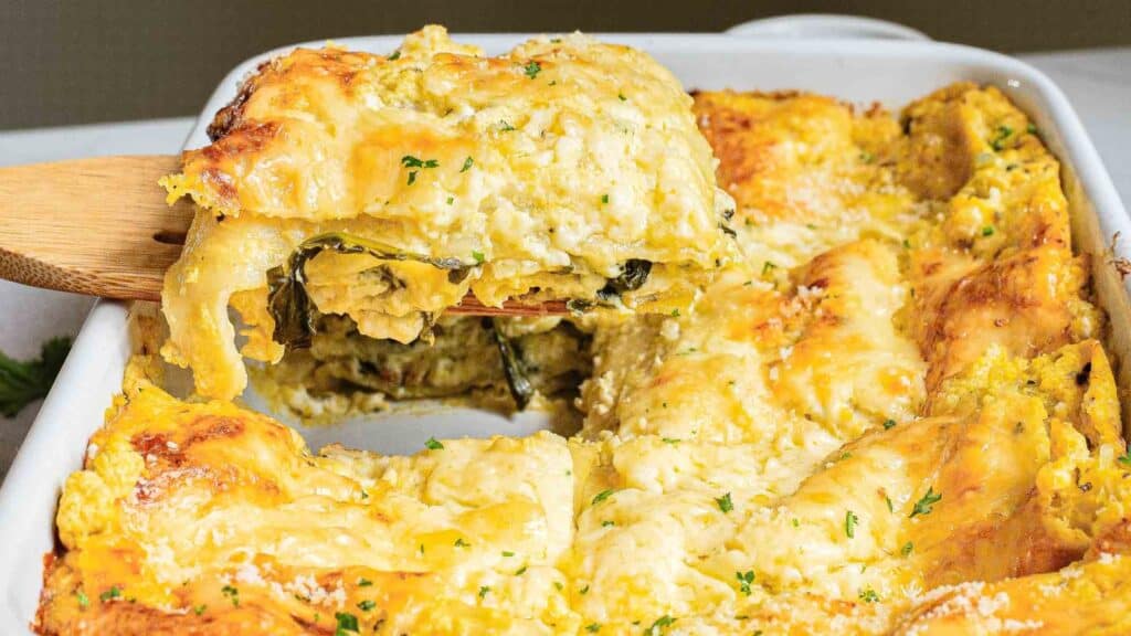 A baked spinach and cheese lasagna, topped with melted cheese, is being served from a rectangular white dish using a wooden spatula.