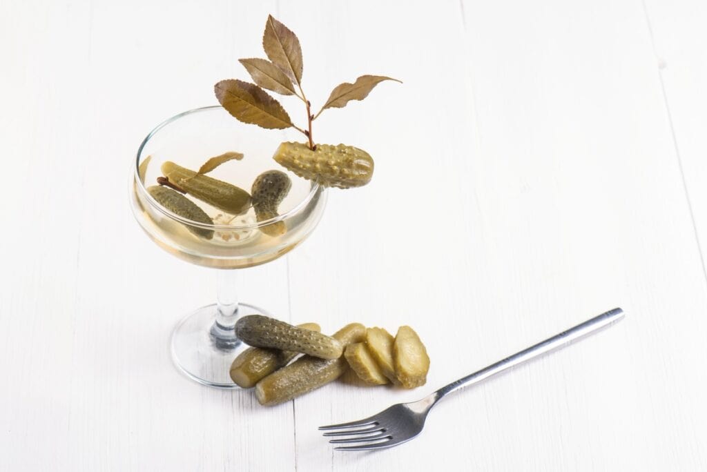 A cocktail glass filled with pickles, decorated with a branch of leaves, next to a fork and more pickles on a white surface.