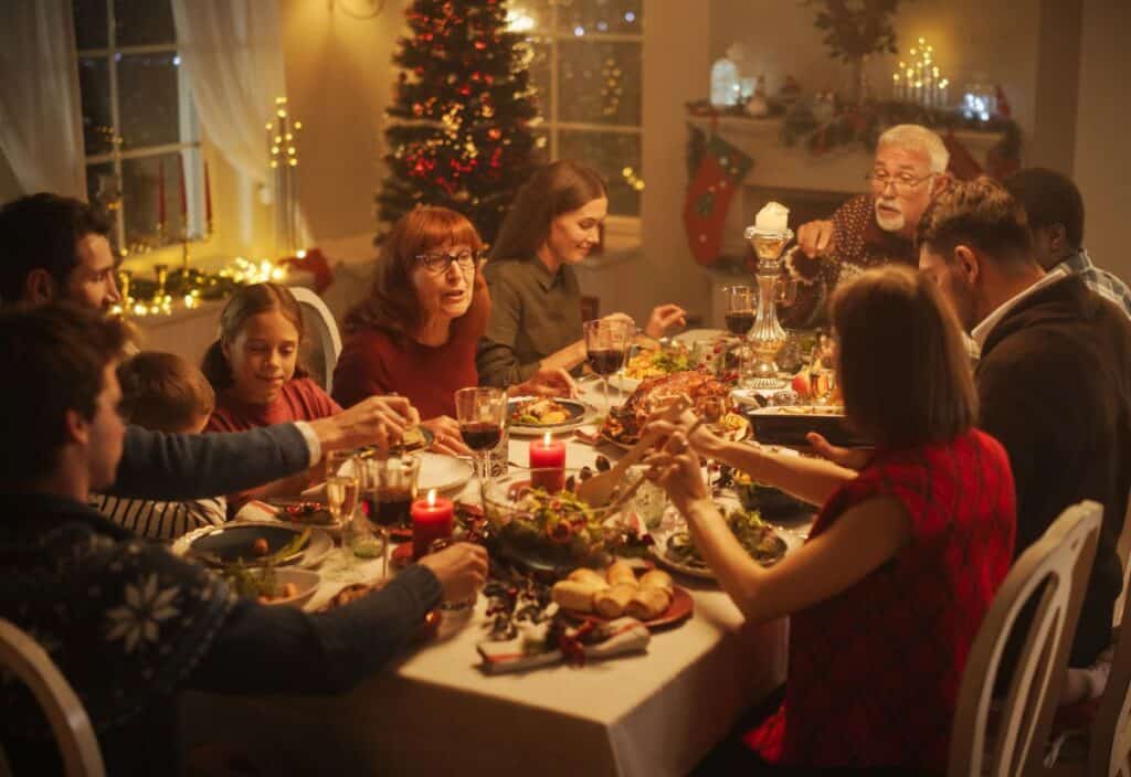 A family gathers around a festive dinner table, sharing a meal. The room is decorated with Christmas lights and a tree, creating a warm holiday atmosphere.