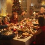 A family gathers around a festive dinner table, sharing a meal. The room is decorated with Christmas lights and a tree, creating a warm holiday atmosphere.