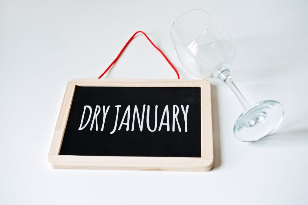 A small blackboard with "Dry January" written on it lies beside a tipped wine glass on a white surface.