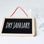 A small blackboard with "Dry January" written on it lies beside a tipped wine glass on a white surface.