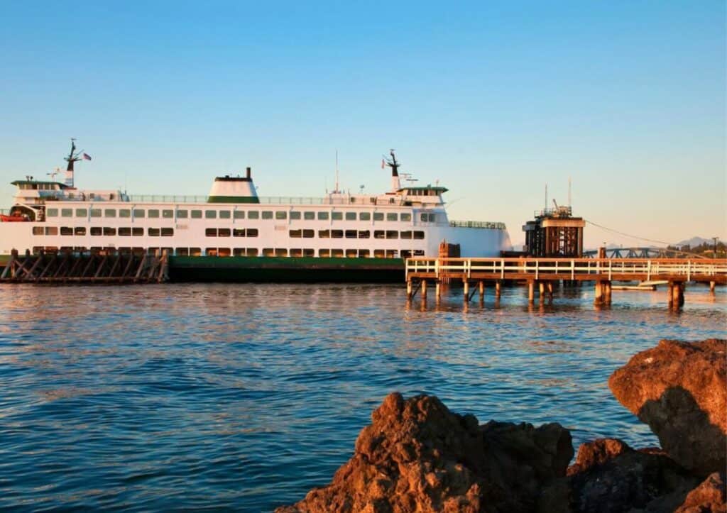 A large ferry is docked at a pier under a clear blue sky, with calm water in the foreground and rocks at the water's edge.