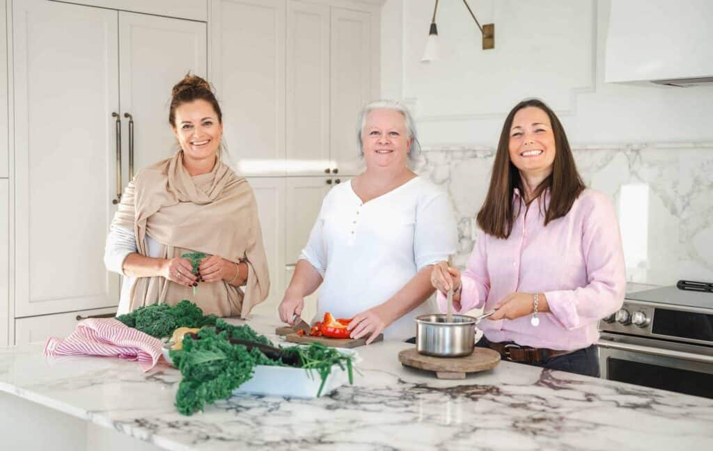 Three women are standing in a kitchen. One is holding greens, another is chopping vegetables, and the third is stirring a pot on the counter.