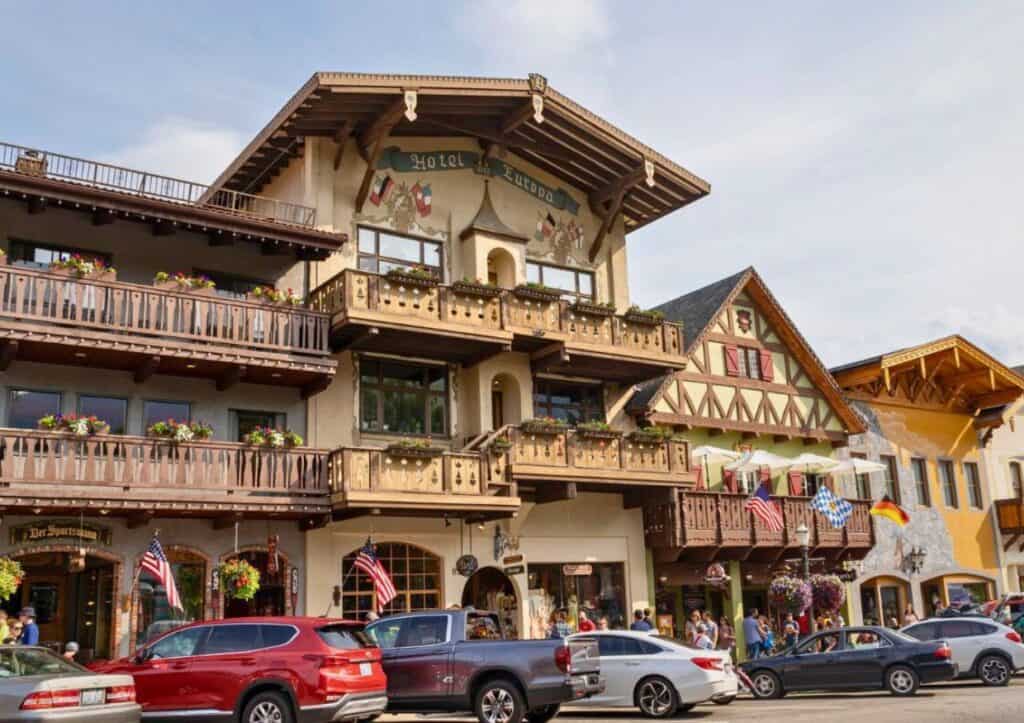 A charming street scene featuring Bavarian-style buildings, cars parked along the road, and people walking by. The architecture includes wooden balconies and decorative elements.