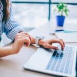 A person in a striped shirt is thoughtfully using a laptop at their desk, surrounded by a small plant and notebook, possibly exploring AI for travel planning to streamline their next adventure.