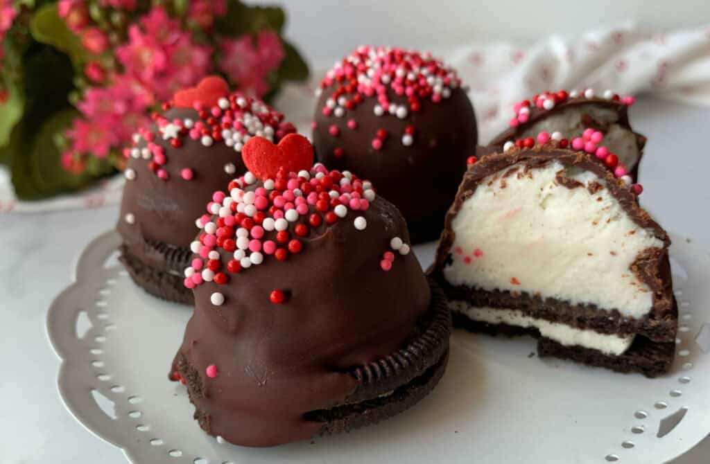 Chocolate-covered desserts with sprinkles and heart decorations on a plate; one is cut in half, revealing a creamy filling. Pink flowers are in the background.