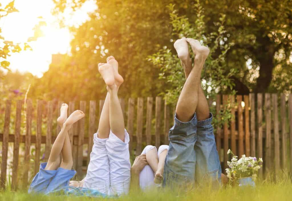 Four people lying on grass with legs raised, wearing casual clothes, next to a wooden fence and flowers, in a sunlit garden setting.