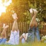 Four people lying on grass with legs raised, wearing casual clothes, next to a wooden fence and flowers, in a sunlit garden setting.